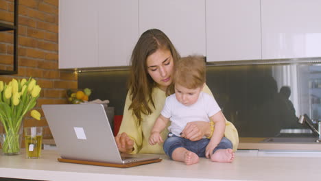 Woman-Holding-His-Baby-Sitting-On-The-Kitchen-Counter-While-Using-The-Laptop