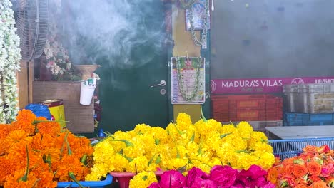 incense burner emits fragrant smoke above vibrant jasmine and marigold garlands in a colorful singapore market stall, captured in bright daylight with a static camera