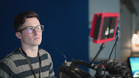 man operating camera in studio environment, focusing on control panel, wearing glasses, looking at camera screen. broadcast professional monitoring equipment, adjusting video settings