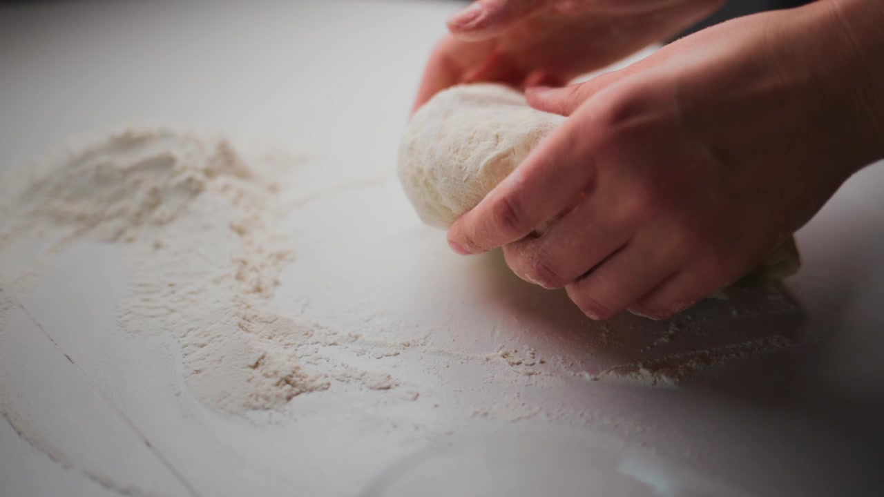 Le mani impastano la pasta bianca su una superficie infarinata. Un primo piano mostra la farina che ricopre l'area di lavoro. La persona si concentra intensamente sulla modellatura dell'impasto. Ideale per cucinare, cuocere al forno o tutorial di ricette.