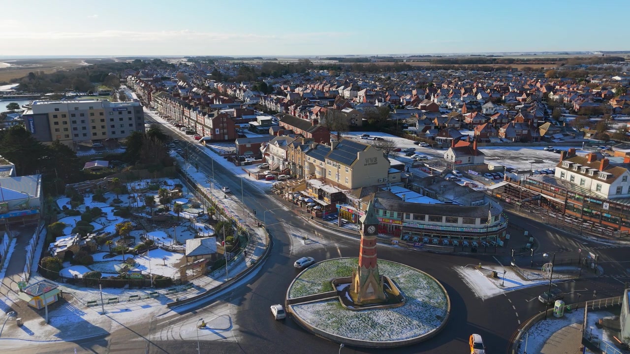 Flygvy fångar ett snötäckt torg med en rondell. Rondellen har ett högt monument i mitten. Snö täcker hustak och vägar under en klar himmel.