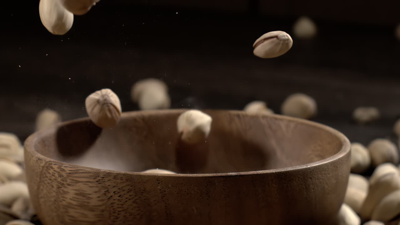 Ciotola di legno piena di pistacchi su tavolo di legno scuro, gustosa frutta secca salata sparsa intorno, concetto di snack salutare e cibo naturale. Scatto con telecamera super slow motion a 1600 fps