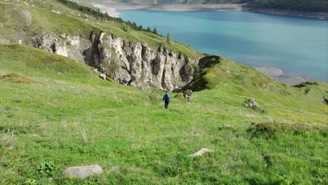 Vista-Aérea-De-Un-Hombre-Caminando-Por-Una-Ladera-Verde-Junto-Al-Lago-Mont-Cenis