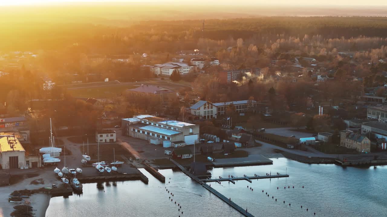 Engure Port glows under golden hour light as sunlit waters and peaceful docks stretch into the harbor, with the coastal town fading into the warm evening haze.
