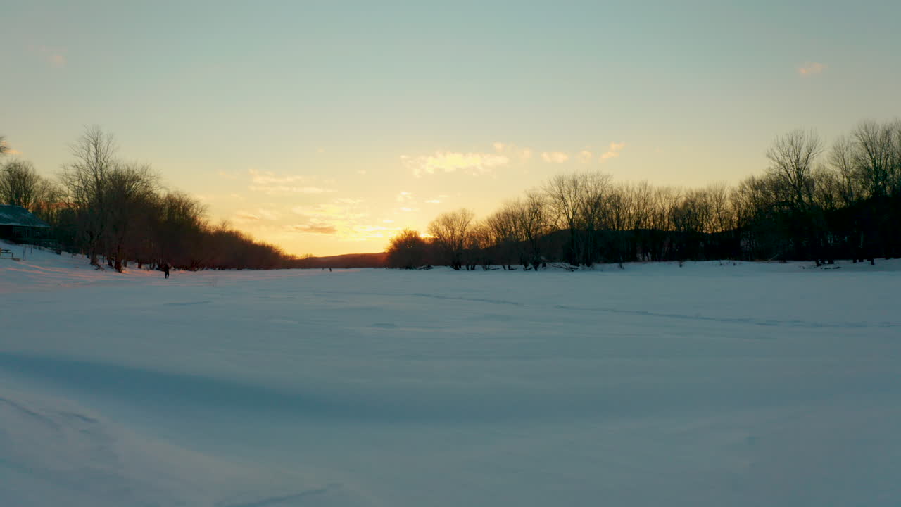 석양에 얼어붙은 눈 덮힌 강을 따라 개를 산책시키는 사람들을 드러내기 위해 다리 아래로 날아가는 공중 전망