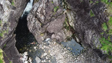 Woman-in-bikini-relaxing-inside-river-pond-in-rocky-canyon-with-wild-surroundings---Hesjedalsfossen-waterfall-Norway---Downward-moving-aerial-close-to-cliff