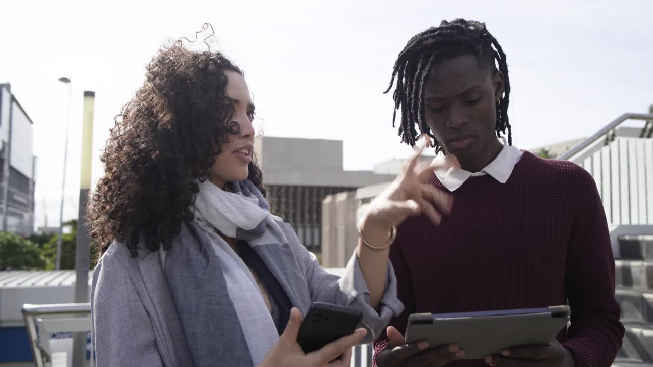 Diverse coworkers pointing phone screen, gesturing and discussing tablet on rooftop terrace