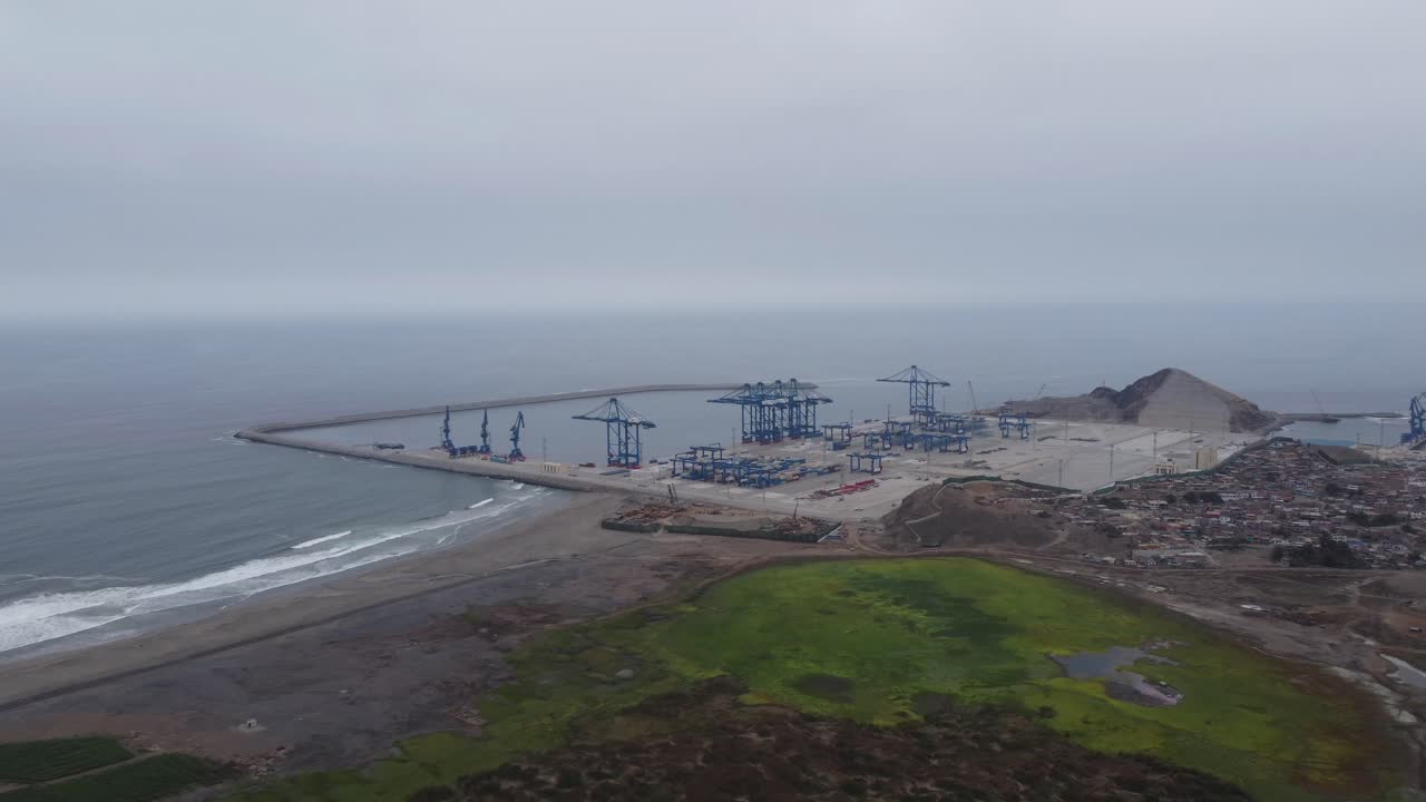 Construction site of a Megaport under construction in the Peruvian city of Chancay. Drone hovers and pans left to right along the coast showing the harbor and many big blue cranes in the distance.