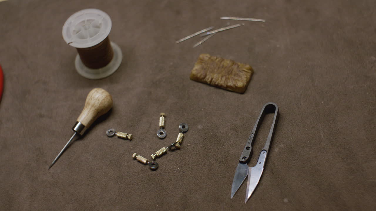 A set of leatherworking tools, including an awl, thread spool, scissors, needles, and brass fasteners, arranged on a brown suede work surface.