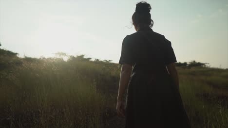 Toma-Cinematográfica-De-Una-Mujer-Joven-Con-Un-Vestido-Negro-Caminando-Por-Un-Campo-De-Hierba-Hacia-El-Sol-Brillante