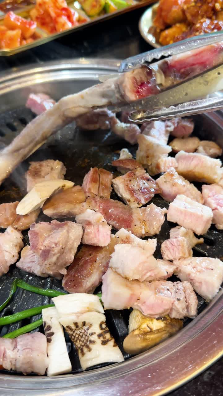 Tangan menggunakan penjepit dan gunting untuk memotong iga babi di atas panggangan meja, pencahayaan restoran yang semarak