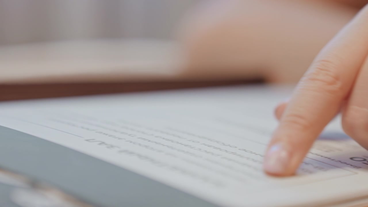 Primo piano di un dito che segue un testo stampato, luce diurna soffusa sulla scrivania dello studio, studente attento che ripassa gli appunti, movimento lento e deliberato, bordo della pagina e sfondo sfocato, focus sulla punta del dito e sulla riga di lettura