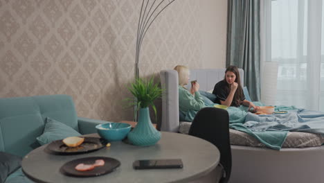 two girls sit comfortably on bed covered with teal blankets, chatting and enjoying snack and coffee in cozy bedroom with soft light, stylish decor, and warm relaxed ambiance near window