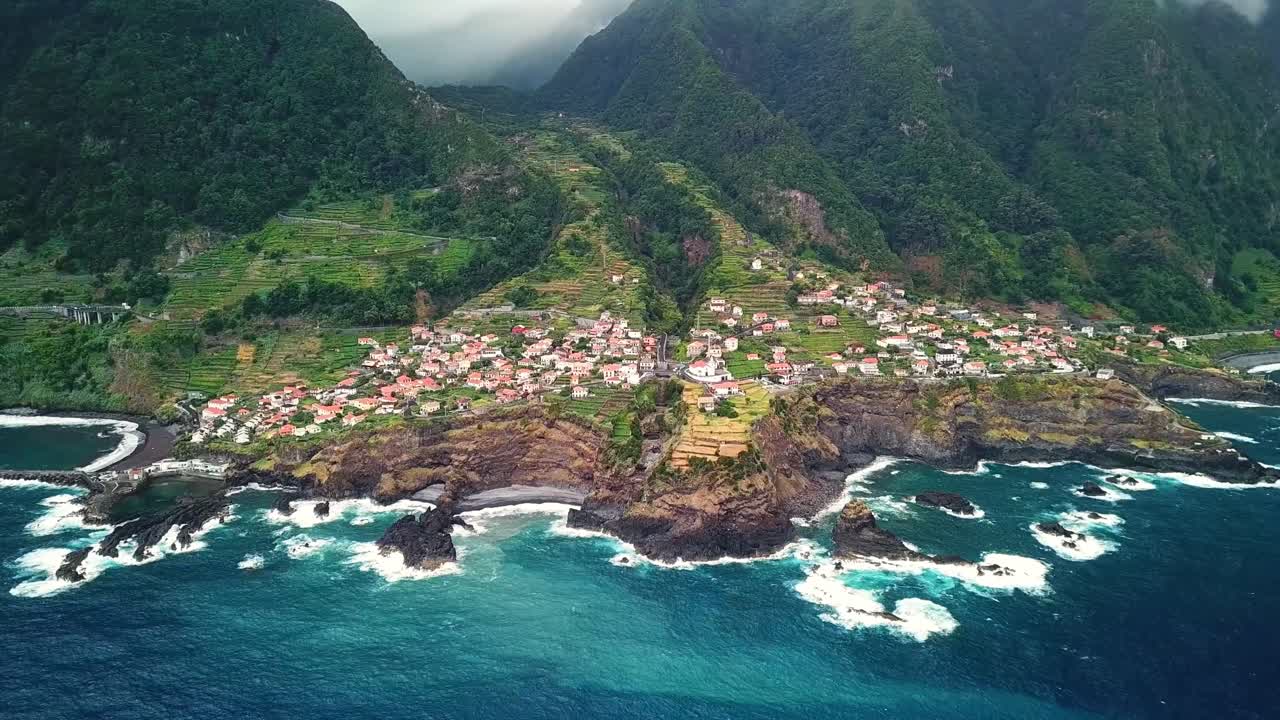 Aerial view of Seixal, Madeira, a picturesque coastal village with terraced green hills, dramatic cliffs, and crashing waves. Vibrant houses and natural beauty highlight the tranquil seascape