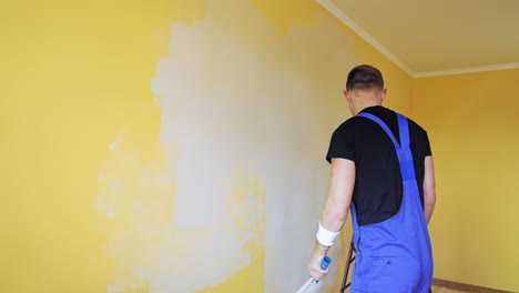 Worker lightening walls in the room. Young builder in blue overalls doing renovation in the apartment with a paint roller.