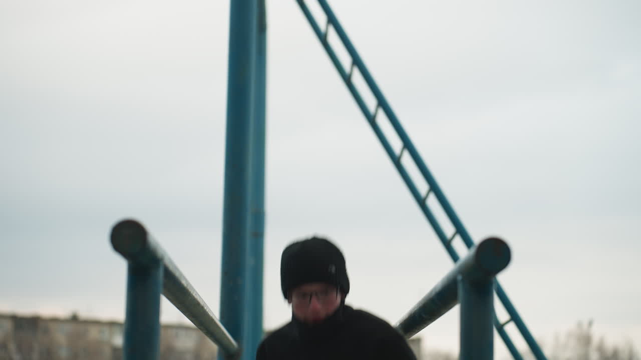Close-up of a young boy finishing a workout on a metal bar, dressed in a black outfit and glasses, after dropping from the bar, he walks away