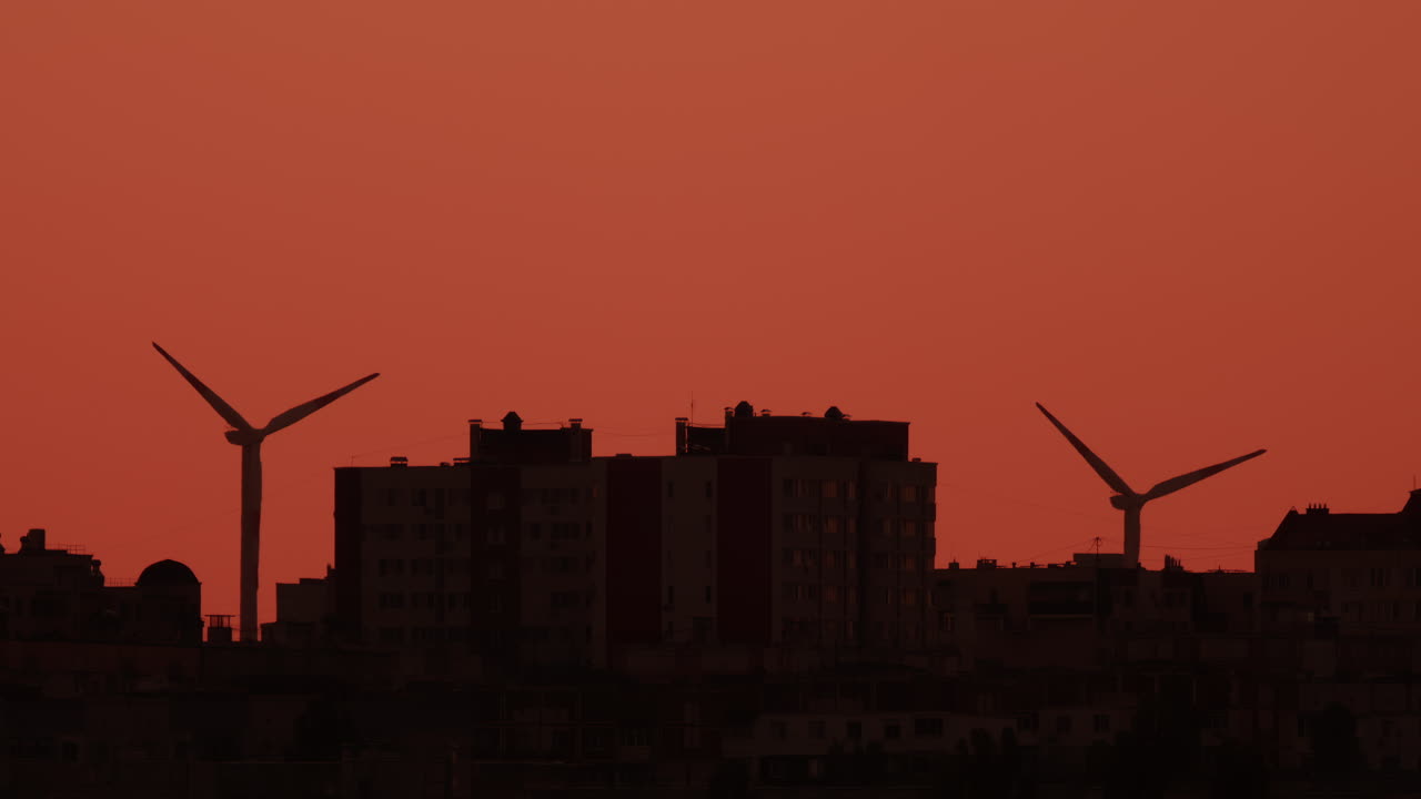 Two wind turbines stand tall near city buildings under an orange sky