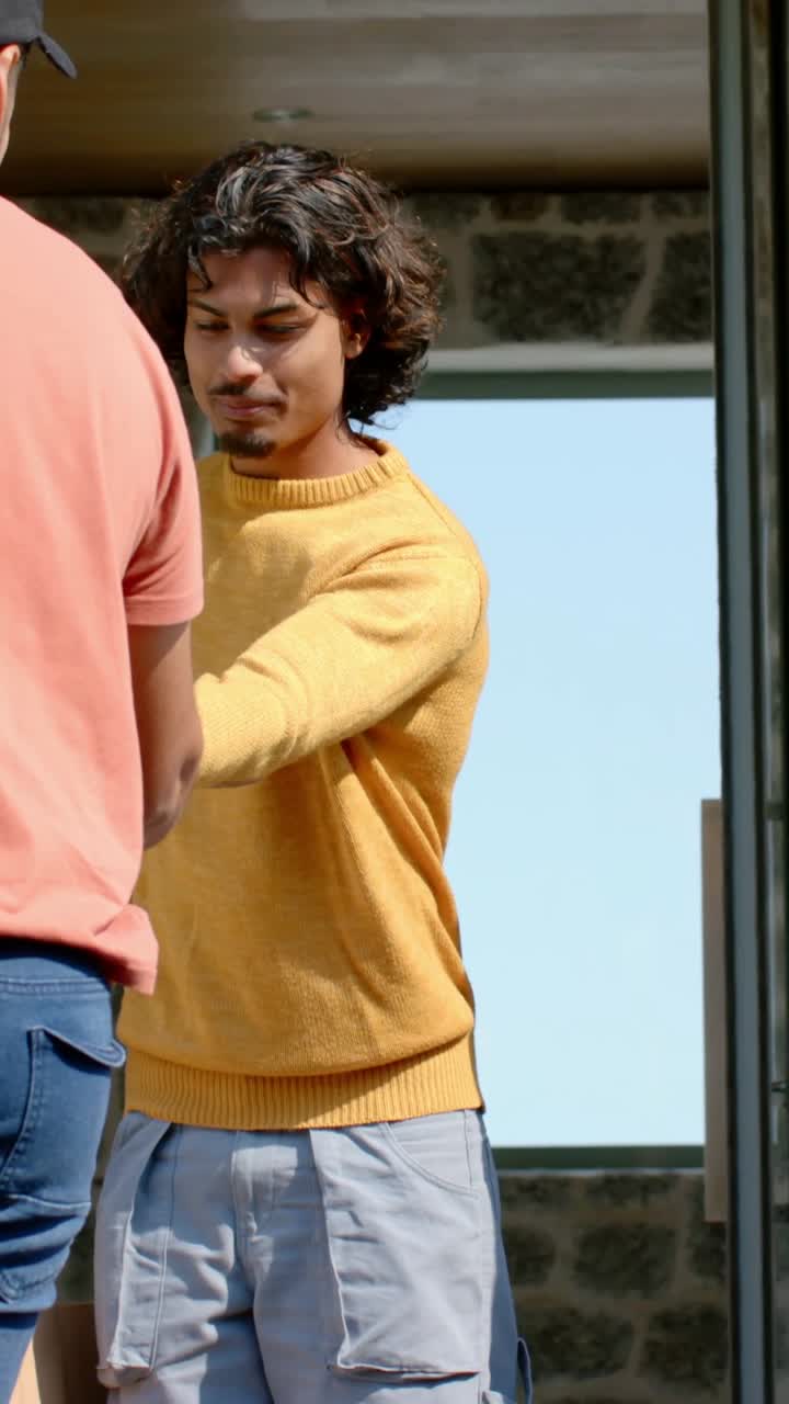 Video verticale: Uomo con maglione giallo che si protende per ritirare un sacchetto dell'ordine alla porta, mentre un amico gli porge un sacchetto rosso.