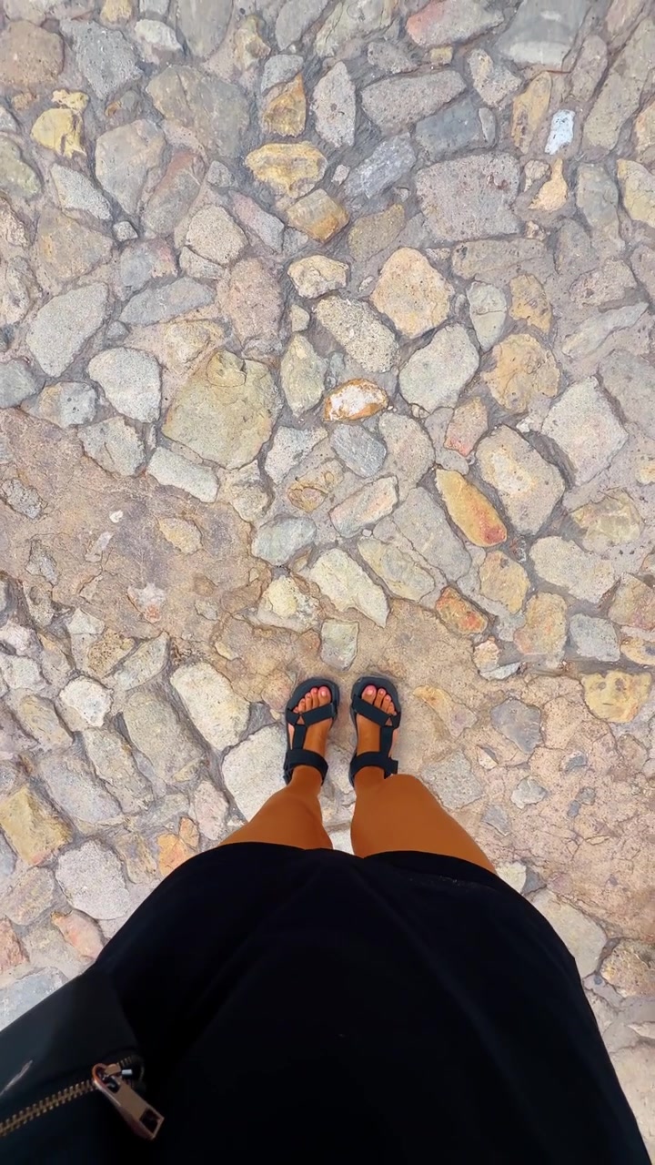 Persona in sandali che cammina su un vecchio selciato di ciottoli. Vista dall'alto di gambe e piedi con sandali neri a fascia che camminano su una pavimentazione in pietra irregolare in una città storica.