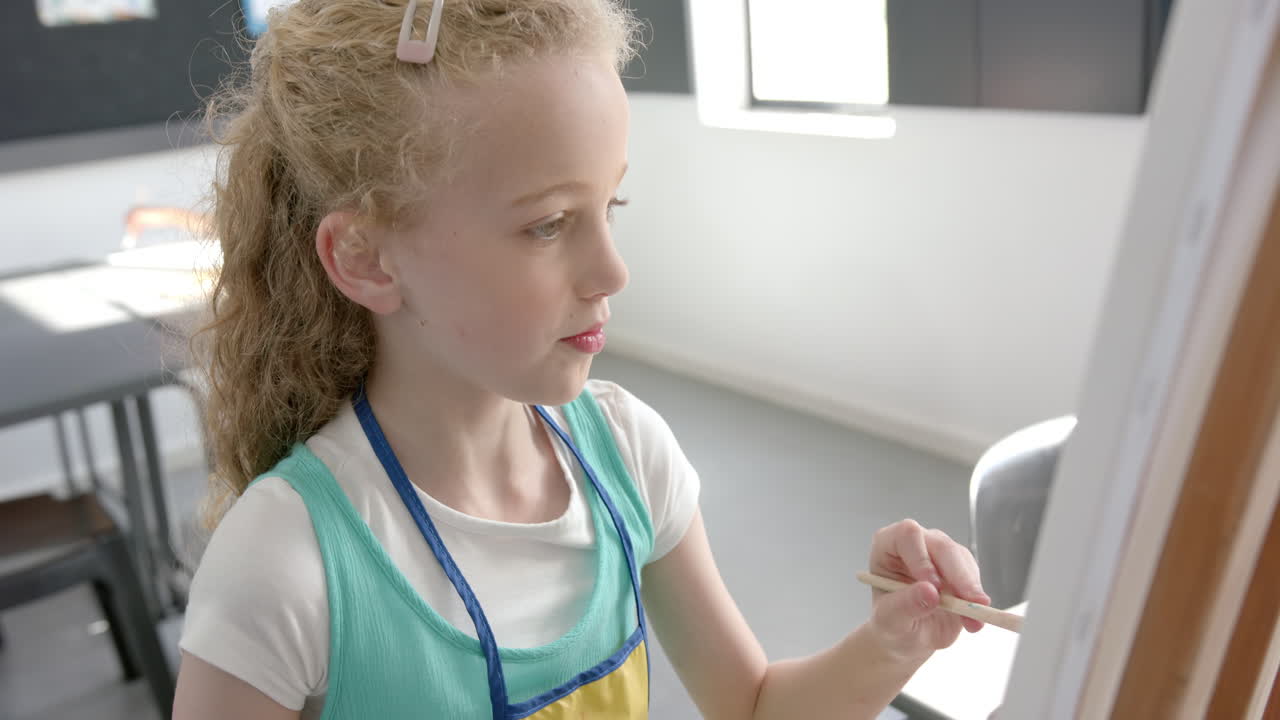 Caucasian girl with blonde hair is painting on a canvas in a classroom at school