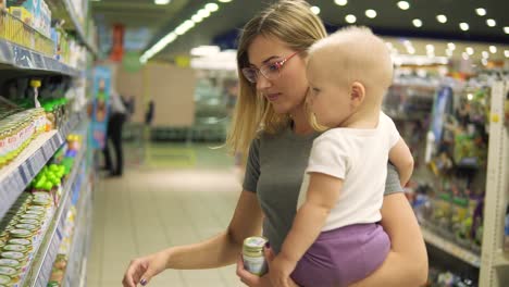 joven madre atractiva con gafas sosteniendo a su hijo en sus brazos mientras elige comida para bebés en los estantes del supermercado