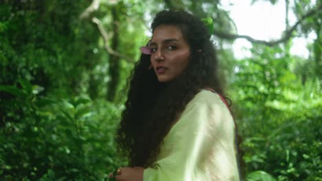 close-up of a woman with long curly hair and a green scrunchie, dressed in traditional clothes. she turns slightly, then faces away in a lush green jungle