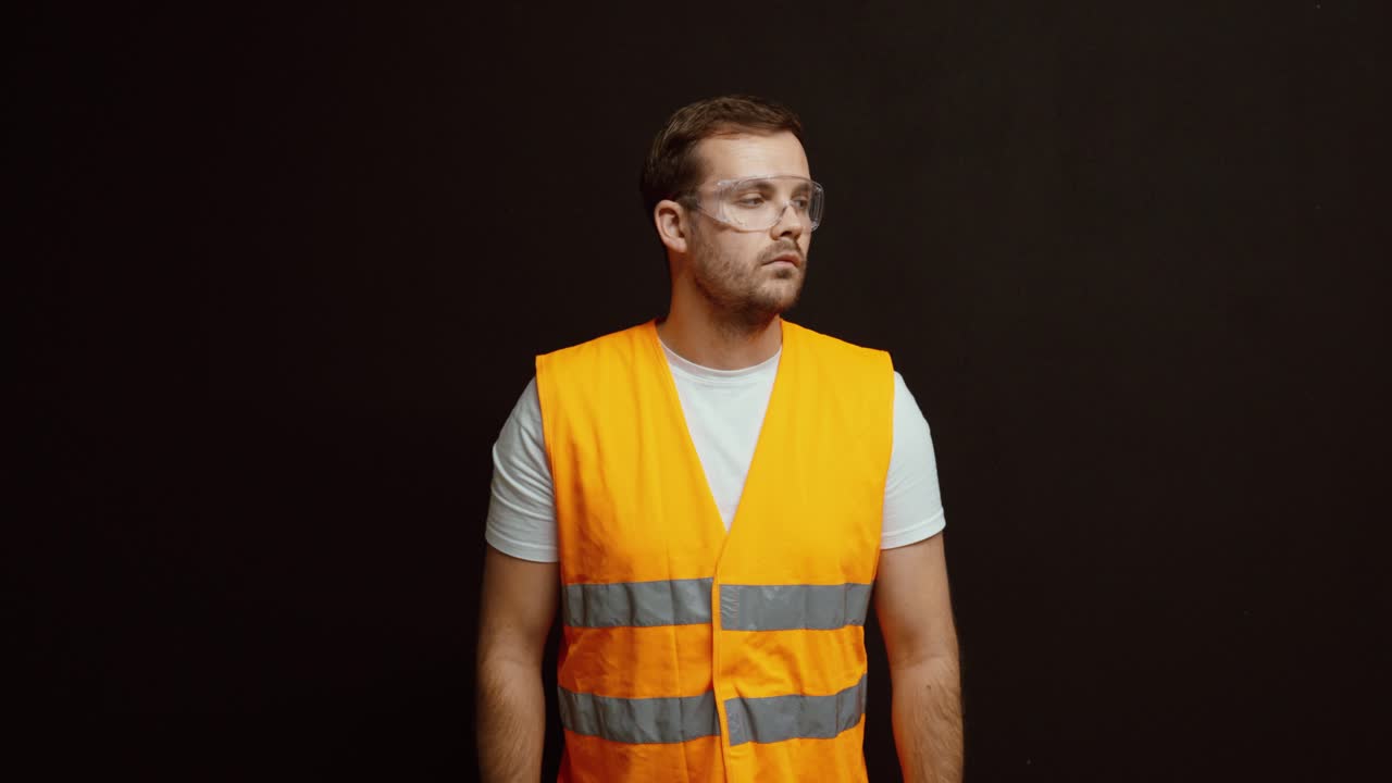 A worker in a high-visibility vest holds and puts on safety goggles, demonstrating proper workplace safety and protection practices