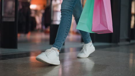 vista de la pierna de una persona con vaqueros y zapatillas de deporte de lona blanca sosteniendo bolsas de compras rosadas y menta mientras camina a través de un centro comercial más allá de una tienda de ropa, con fondo borroso de pantallas minoristas