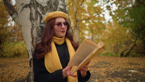 student in solitude reads through yellow covered book with warm smile, seated alone in nature, wearing yellow beret and yellow muffler, leaning against birch tree surrounded by autumn leaves