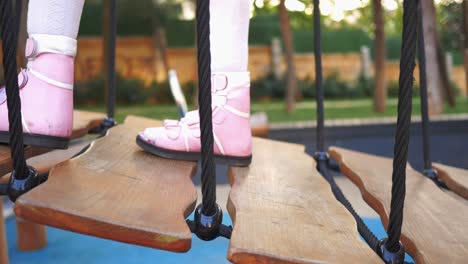 una niña pequeña con zapatos rosados camina por un puente de madera en un patio de recreo