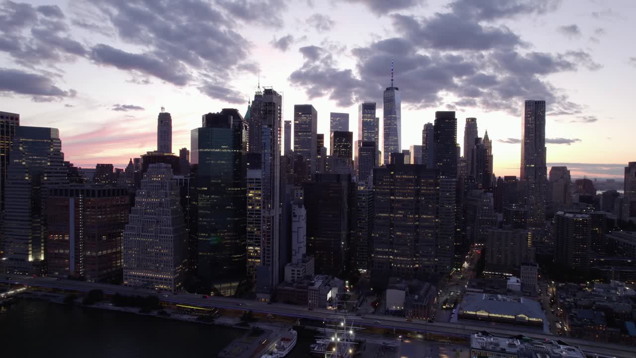 Aerial view away from illuminated skyscrapers in the Manhattan skyline, colorful evening sky in New York, USA - pull back, drone shot