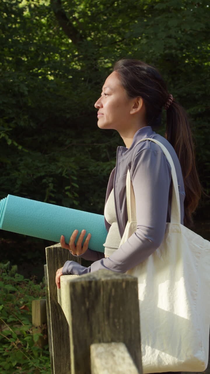 Video verticale di donna che indossa abbigliamento sportivo, con tappetino da yoga, in piedi su un ponte nella foresta, che si guarda intorno godendosi la pace e la bellezza della natura. Ripreso in tempo reale 1.