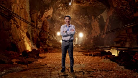 full body shot captures of an asian male professional worker standing in underground mine tunnel, his broad smile at the camera and cross his arm over his chest