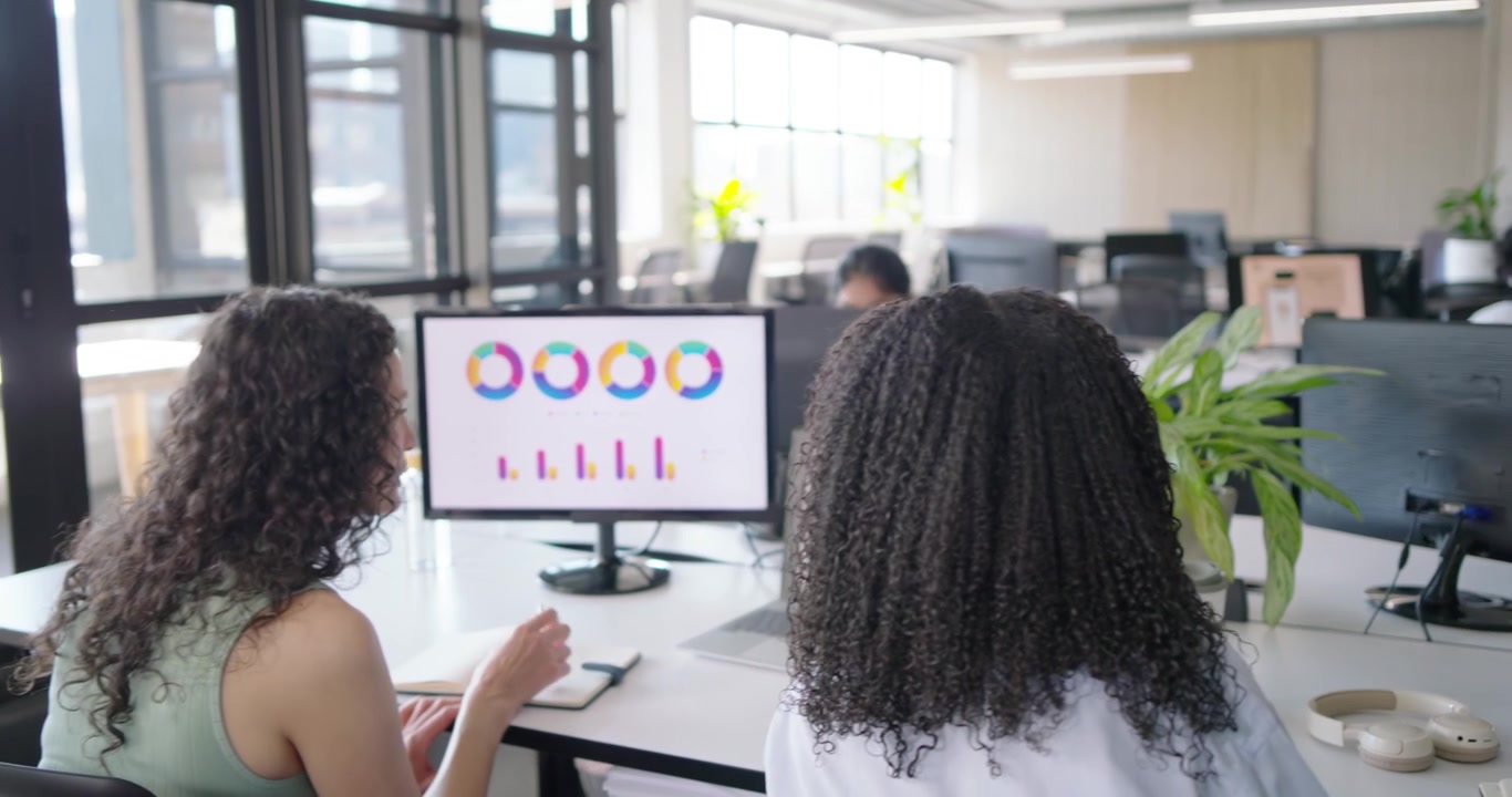 Diverse female coworkers discussing and analyzing monitor charts while making plans at desk laptops