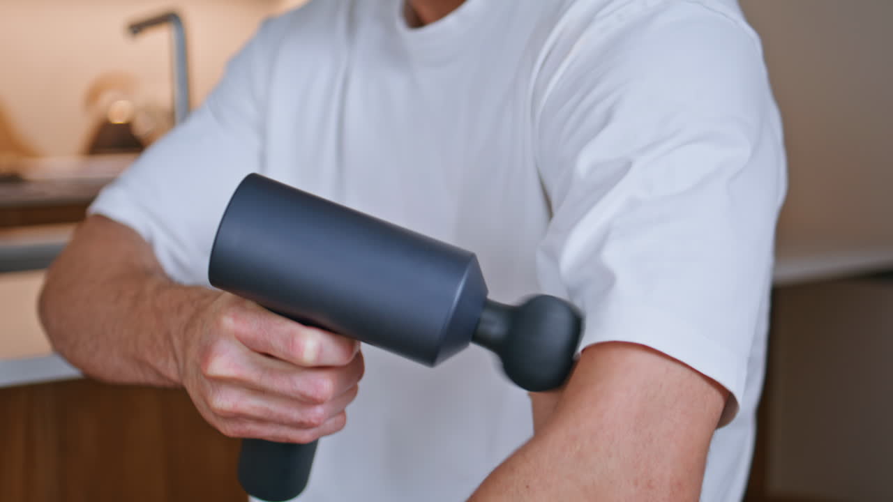 Mãos segurando uma pistola de massagem aliviando a dor muscular no braço em close-up na cozinha de casa