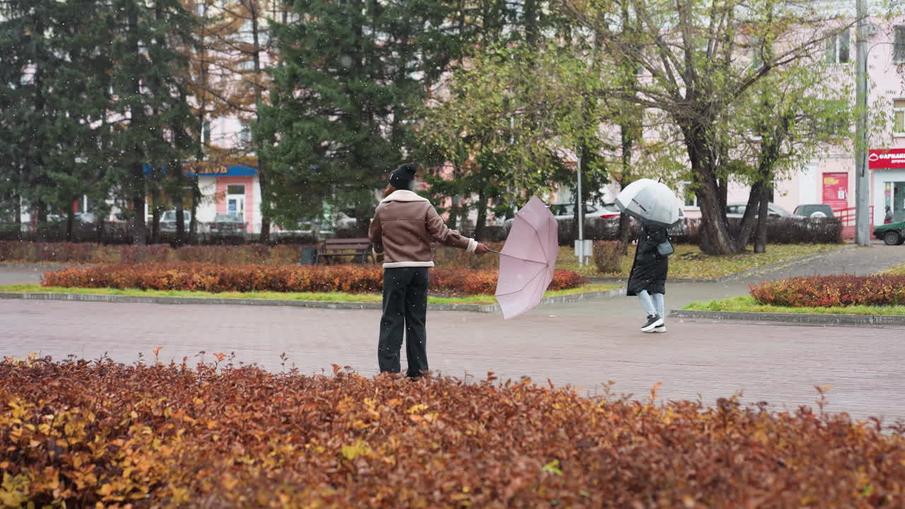Happy lady holding umbrella, wearing knit cap, brown shearling jacket, black trousers, happily turning around in light snowfall, enjoying winter day, spreading positive energy in cold weather