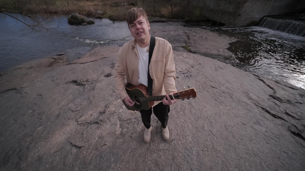 바위의 배경에 산의 강변에 앉아 기타를 연주하는 젊은이. 자유의 개념, 휴식 장소.