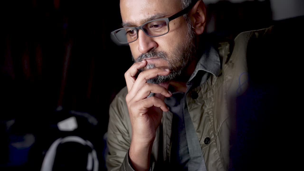 A thoughtful South Asian man wearing glasses sits in a dim room, lost in thought