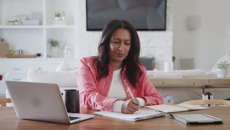 Middle-aged-woman-sitting-at-a-table-at-home-making-notes-in-a-notepad,-front-view,-panning-shot