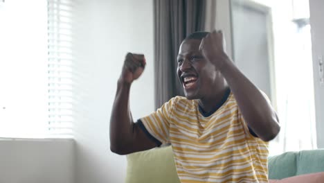 happy african american man watching match and celebrating at home, slow motion