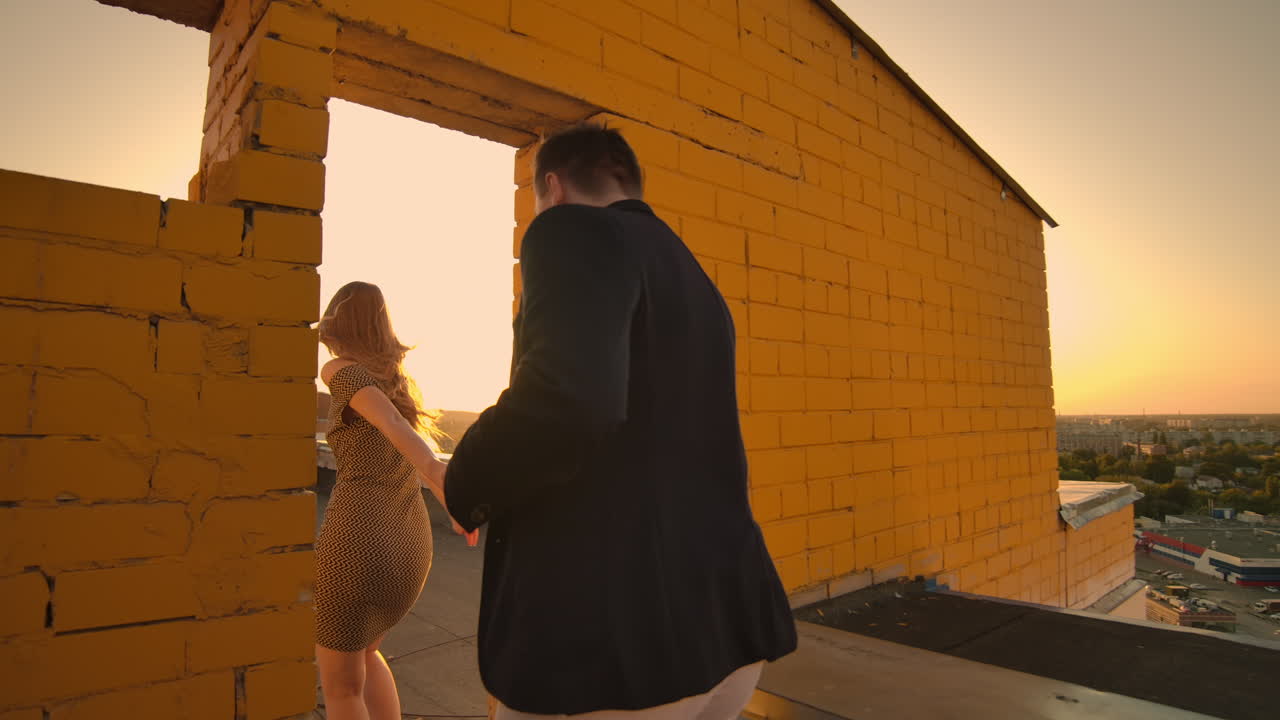 웃는 귀여운 연인들은 도시의 배경에서 해가 지는 건물의 지붕에서 달리고 가장자리에 서서 포옹합니다.