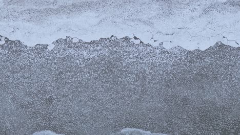 edge of a frozen lake with ice meeting a snow-covered shore, bird's eye view, in daylight