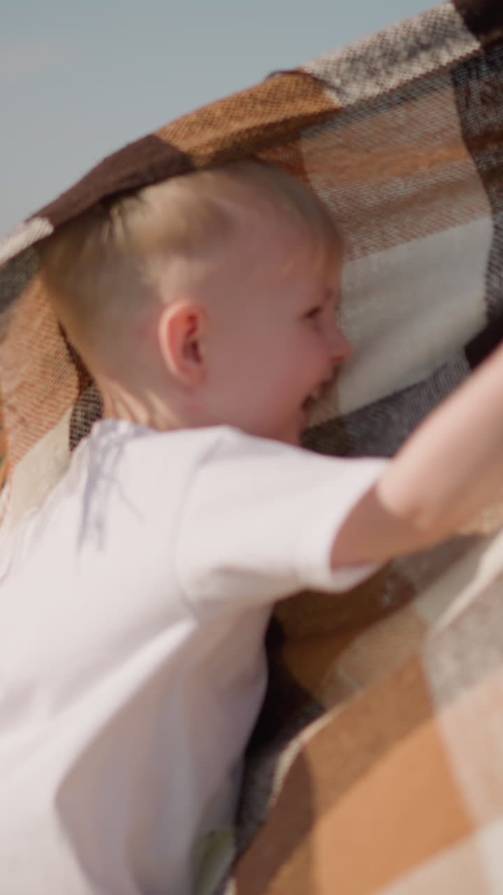 A young boy in a white shirt playfully hits a flowing plaid scarf in a grassy field. A woman and another boy are in the background, adding to the joyful and carefree atmosphere
