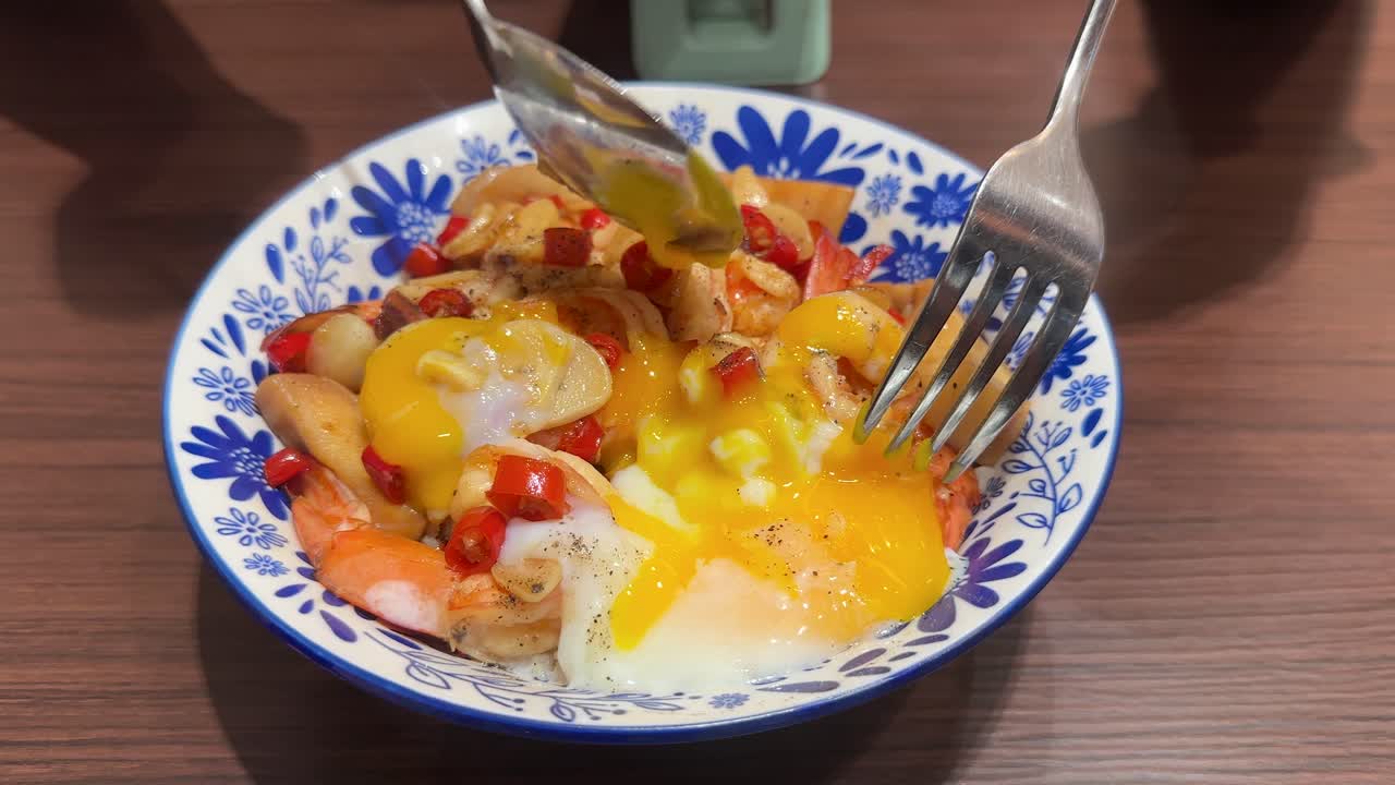 A spoon and fork break and mix a soft-boiled egg into a colorful shrimp rice bowl with chili and garlic, under bright indoor lighting