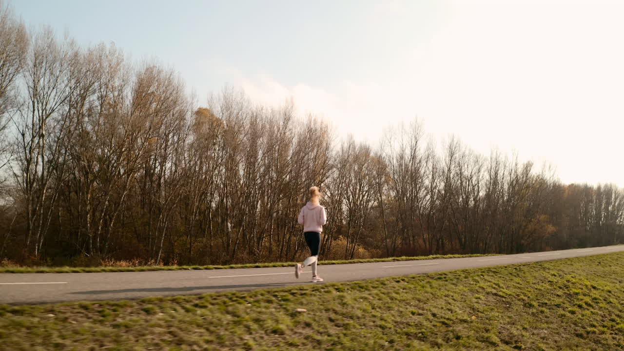 Tracking aerial view of a slim young woman running on a cycling path with autumn trees in the background - stock video