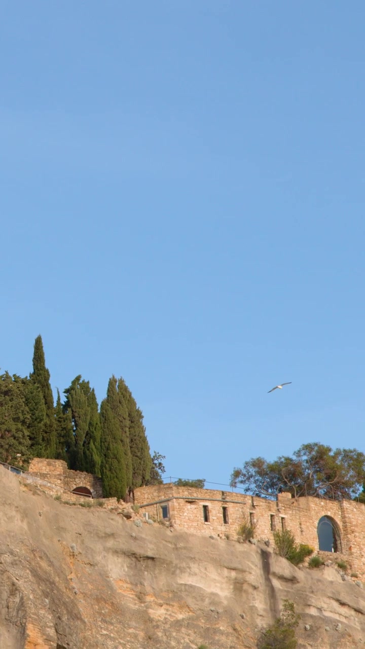 Un'inquadratura ampia e statica di un antico castello in pietra in cima a una scogliera rocciosa sotto cieli azzurri.