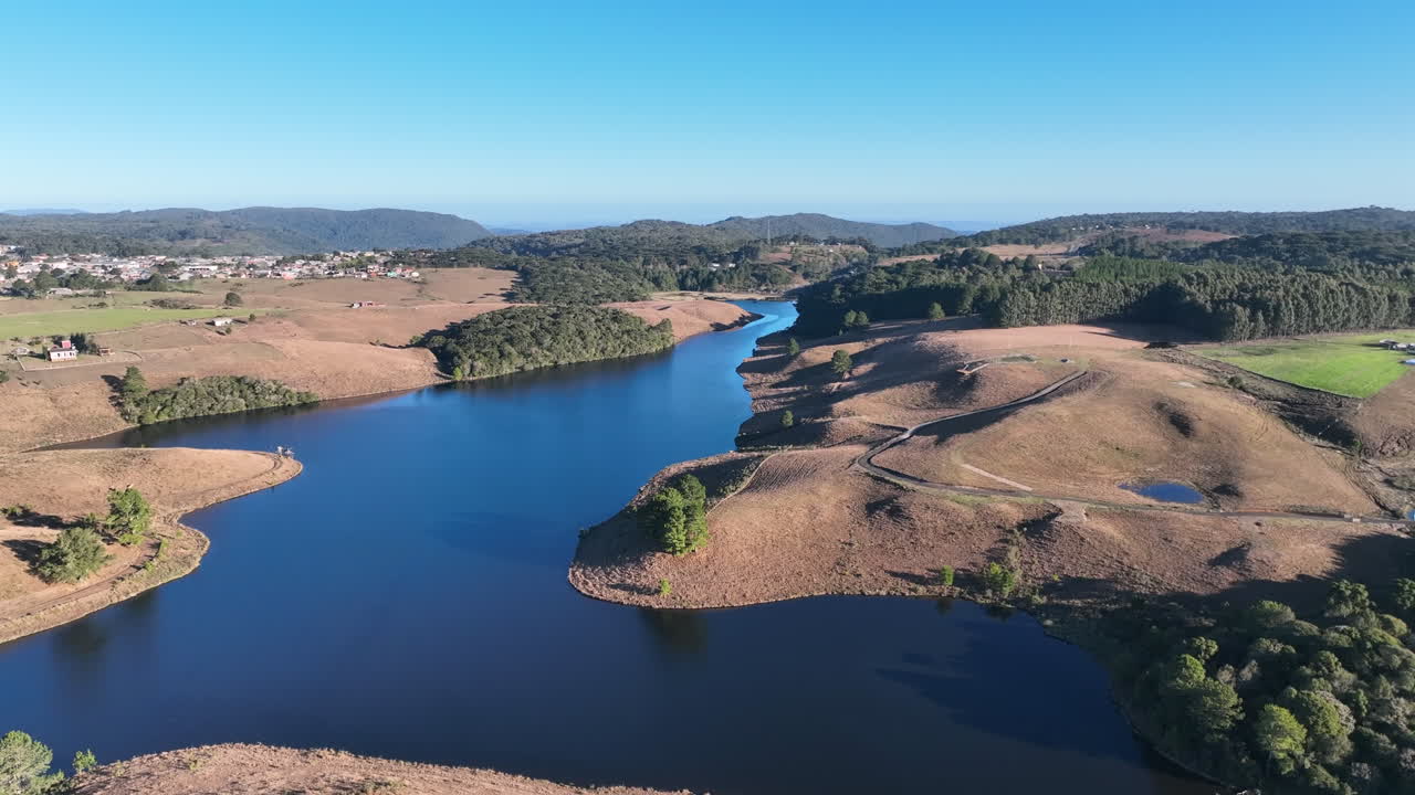 Volo cinematografico ad alta quota con drone e inclinazione cardanica verso il basso su un incontaminato bacino blu. Mostra colline dorate invernali e una città distante sotto la luce della sera in Brasile