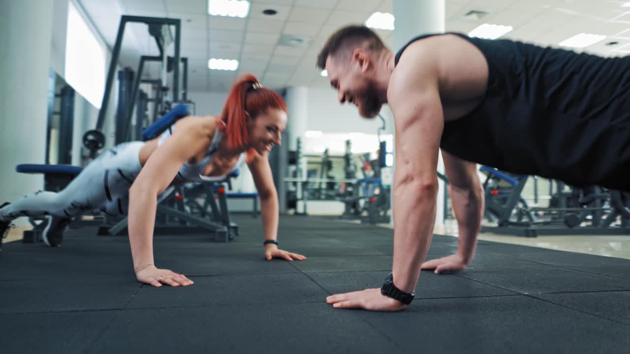 Smiling couple of sportsmen does the push-ups in modern gym. Attractive woman and muscular man training together while doing push ups and clapping by palm in fitness club.