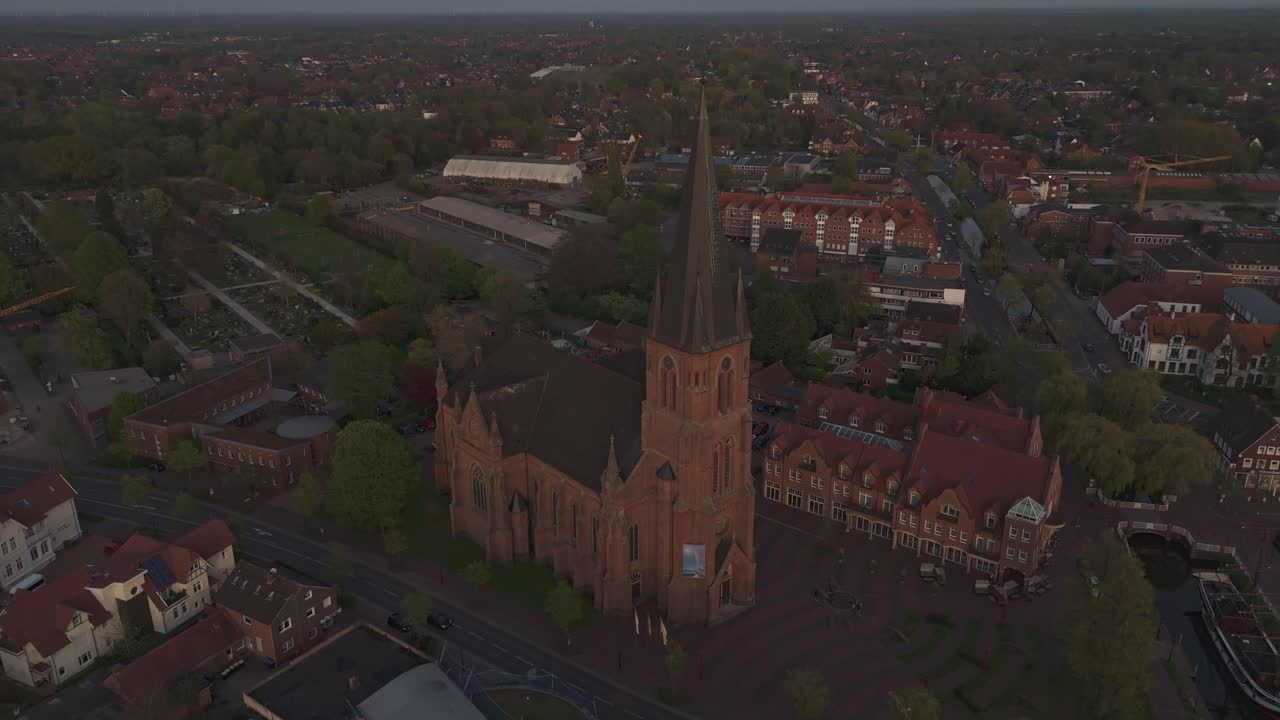 Orbiting drone shot of the red-brick St. Antonius Church in Papenburg, Germany, during golden hour. Surrounded by rooftops, trees and evening light. Historic architecture in a peaceful townscape.