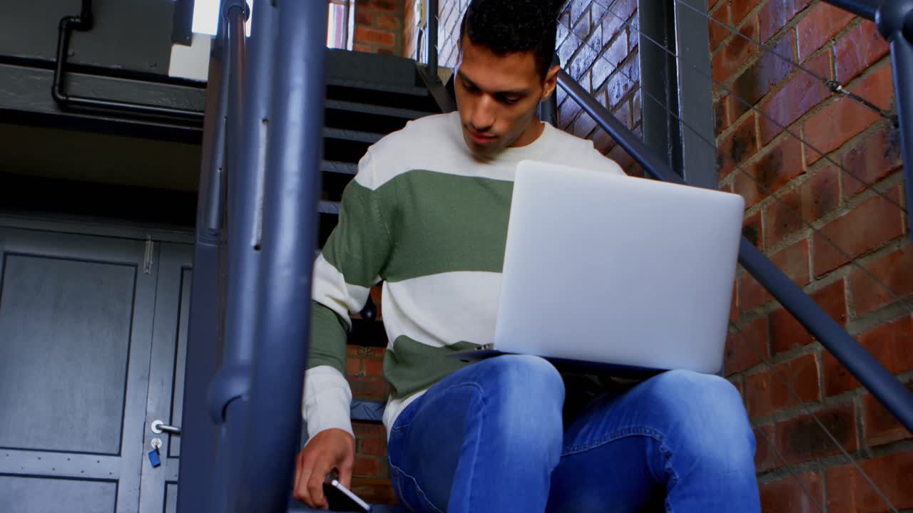 Man talking on mobile while using laptop on stair 4k